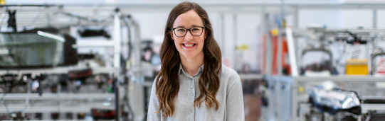 Foto einer lächelnden Frau mit langen, welligen Haaren und Brille. Sie trägt eine helle Bluse und steht vor einem unscharfen Hintergrund, der eine industrielle Umgebung mit verschiedenen Maschinen und Ausrüstungen zeigt. Die Frau blickt direkt in die Kamera.