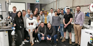 Foto einer Gruppe von Menschen in einer industriellen oder labortechnischen Umgebung. Die Gruppe besteht aus sechzehn Personen unterschiedlichen Alters, die lächelnd in die Kamera blicken. Die meisten stehen, während zwei vorne in der Hocke sind. Die Personen tragen eine Mischung aus Freizeitkleidung und Arbeitskleidung. Im Hintergrund sind industrielle Geräte, Leitungen und Schläuche zu sehen, was auf eine technische oder wissenschaftliche Arbeitsumgebung hindeutet. Die Wände sind grau und es gibt mehrere Türen und Fenster im Hintergrund.