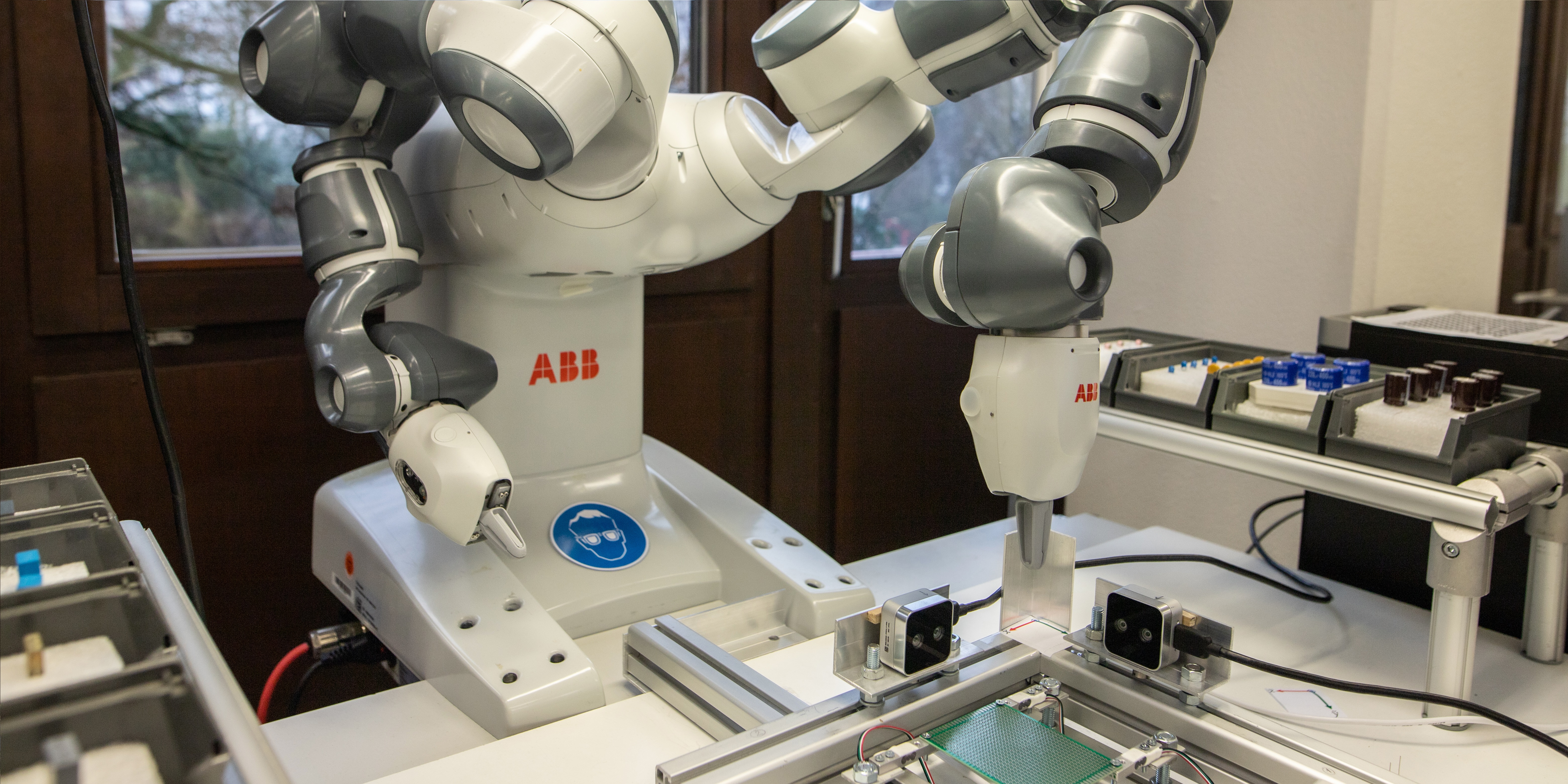 Foto eines ABB-Roboterarms, der in einer industriellen Umgebung arbeitet. Der Roboterarm hat mehrere Gelenke mit grauen und weißen Segmenten. Er bearbeitet präzise eine elektronische Platine, die auf einer Arbeitsfläche vor ihm liegt. Im Hintergrund sind weitere Werkzeuge und Bauteile auf einem Tisch zu sehen. Die Umgebung ist durch große Fenster im Hintergrund gut beleuchtet.