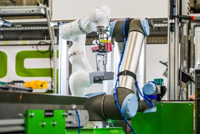 Industrieroboter in einer Fertigungszelle: Ein weißer und ein grauer kollaborierender Roboterarm arbeiten gemeinsam an einem Bauteil. Umgebende Maschinen- und Sicherheitselemente sind sichtbar.