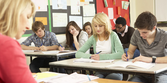 Pupils sit in a class and write in notebooks sitting in front of them a teacher at a desk.