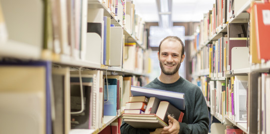 Ein Student steht in der Bibliothek im Gang und hat Bücher auf dem Arm.