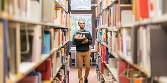 Student in der Bibliothek Studierender läuft an den Bücherregalen in der Bibliothek entlang.